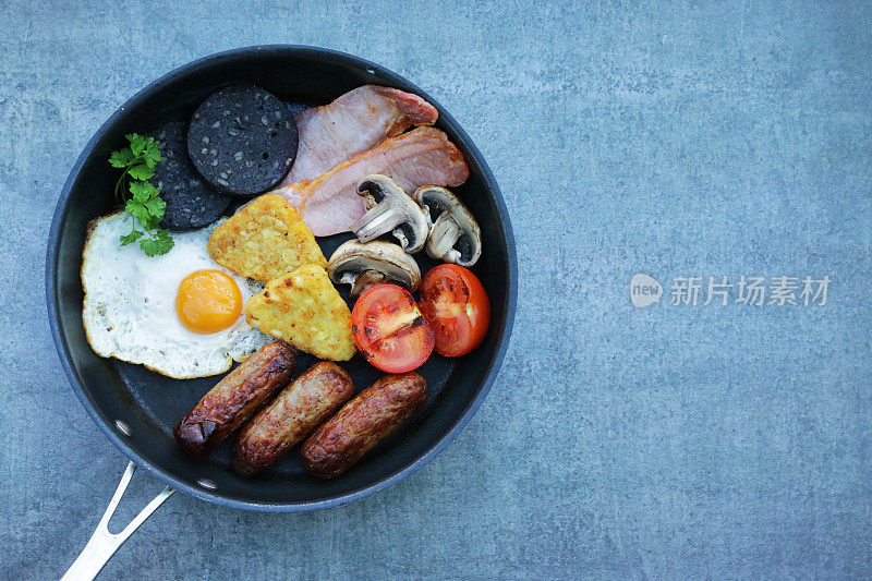 不粘锅的图像包含了完整的英国早餐的元素，煮猪肉香肠，土豆煎饼，西红柿，蘑菇片，培根片，煎蛋和黑布丁(血肠)，灰色的背景，俯瞰