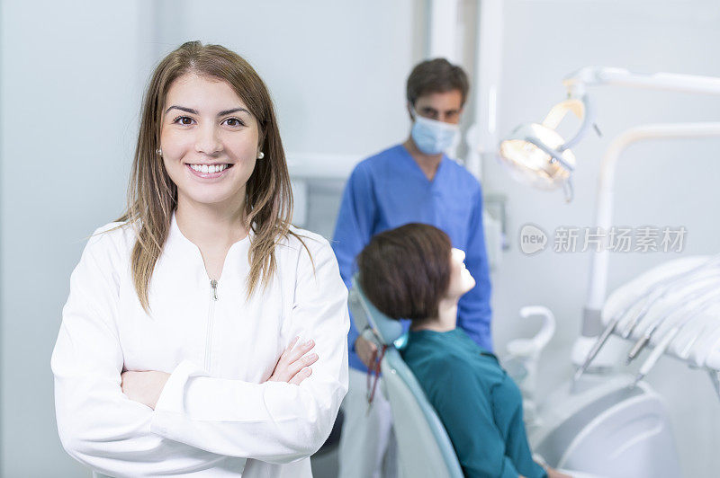 在牙科诊所里，微笑的牙医双臂交叉，看着镜头，背景是病人和助手