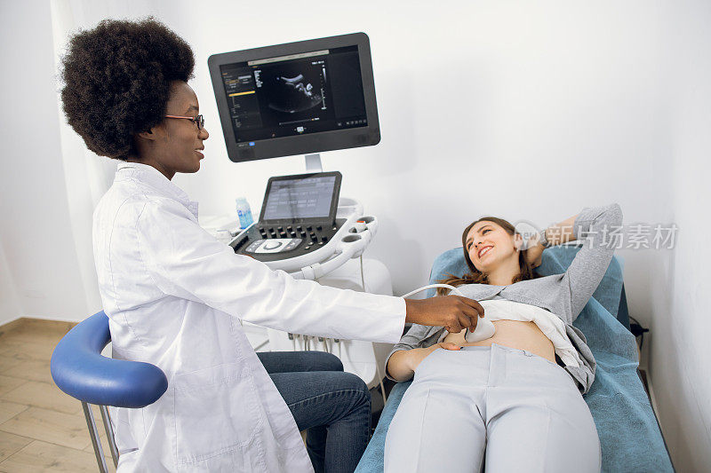 非洲年轻女医生在临床上使用超声波机，为欧洲年轻女患者做腹部超声检查。超声波扫描仪。超声。