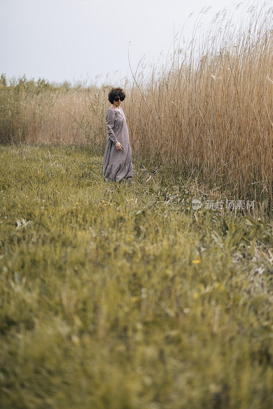 重新连接与自然:年轻的女人与卷发站在一个杂草丛生的田野，穿着长裙