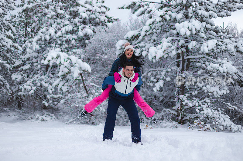 年轻的男朋友背着他的女朋友在雪地里玩，周围是雪树