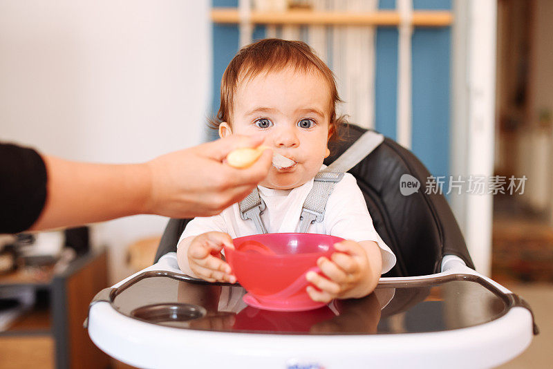 微笑的8个月大男婴坐在家里的高椅子上，妈妈用勺子给他喂固体食物