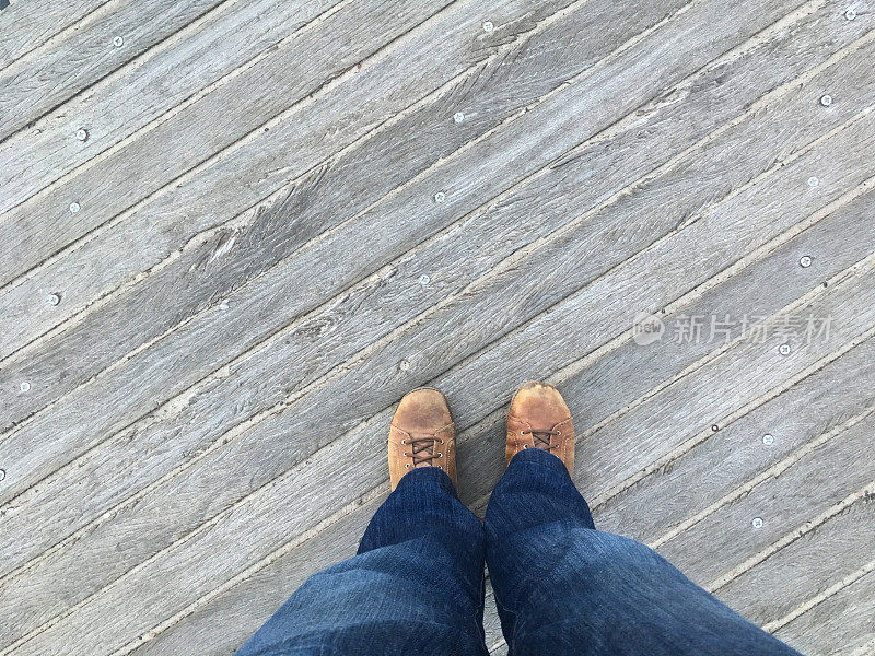 站在纽约布鲁克林科尼岛的木板路上，从脚向下看。低头看着棕色的鞋子穿着粗布牛仔裤，站在外面的木码头上。