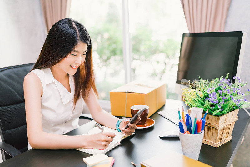 年轻可爱的亚洲小企业主在家办公，用手机打电话，在笔记本上写确认采购订单。收入计划，随意的首席执行官，或创造性的电话营销概念