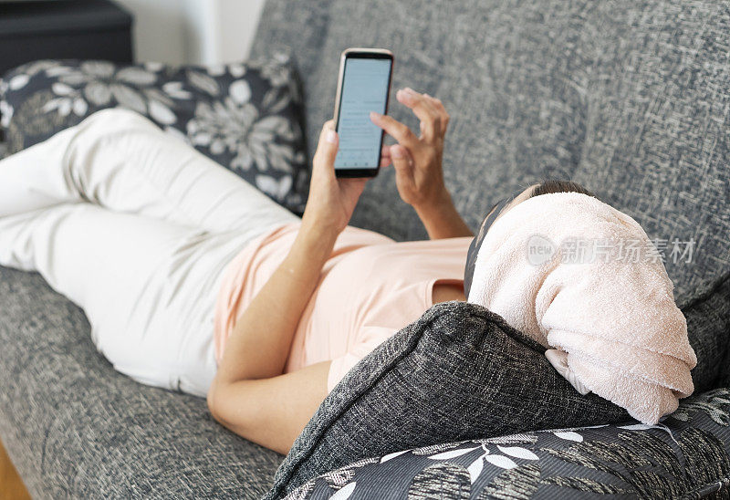 年长妇女带着面膜躺在沙发上，在家里使用手机