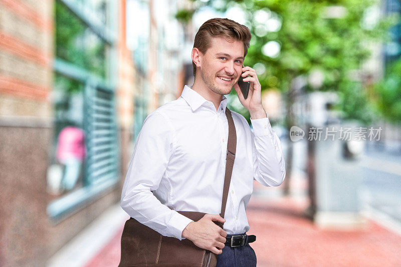 背景模糊的年轻男性商人站在户外，背着肩包，拿着包，使用手机