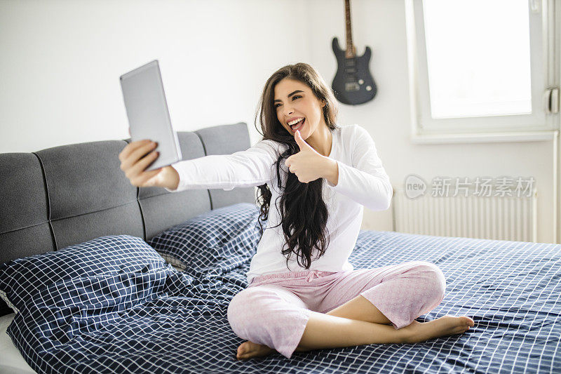 年轻女子在她的床上醒来，她微笑着，并在数字平板电脑上进行视频通话。年轻漂亮的女人在床上醒来，休息得很好。女人醒来后在床上的数字平板电脑上有缩放呼叫。健康的生活方式。健康的概念