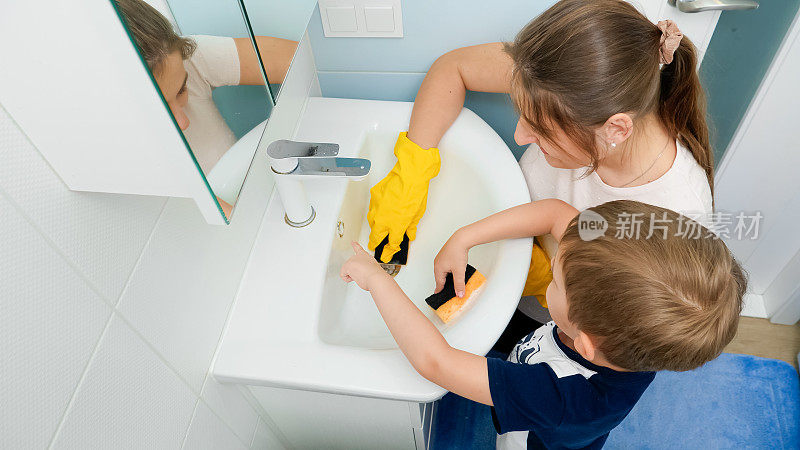 快乐微笑的小男孩和妈妈戴着橡胶手套，在海绵上涂抹清洁剂和清洁剂，洗浴室的水槽。小男孩帮助父母做家务