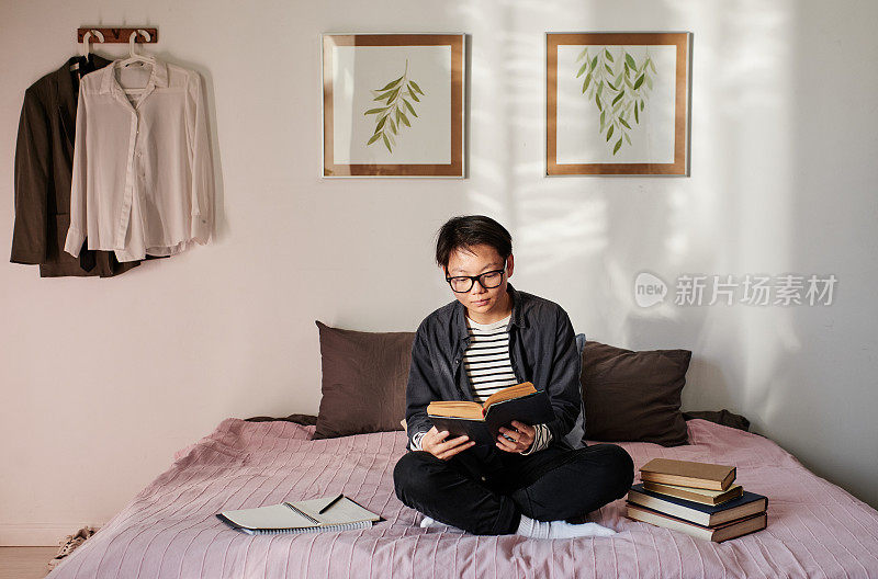 认真的年轻亚洲学生，戴着眼镜，盘腿坐在床上，考试前阅读文献