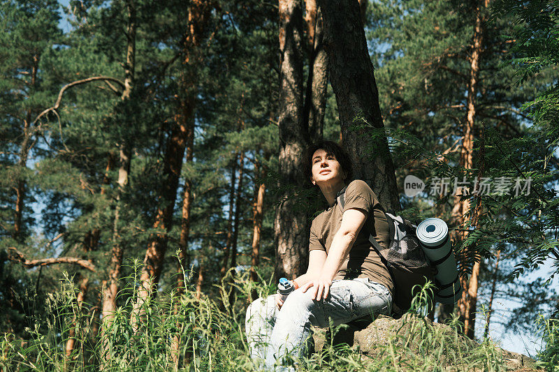 图为一名女背包客在森林中徒步旅行时在石头上休息