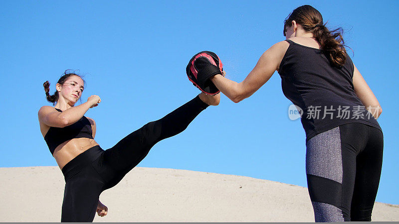 夏天，在烈日炎炎、蓝天下、荒芜的海滩上，两名身穿黑色健身服的年轻健美女子正穿着一双运动鞋，训练打架。慢动作