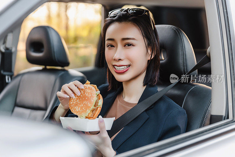 亚洲女人在车里休息，吃汉堡。一个年轻成功快乐的女人正在开车。