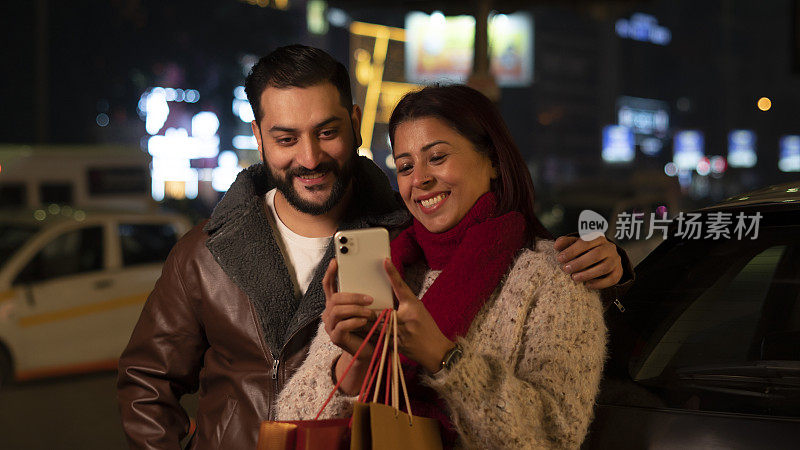 幸福的年轻夫妇晚上在城市购物，使用智能手机