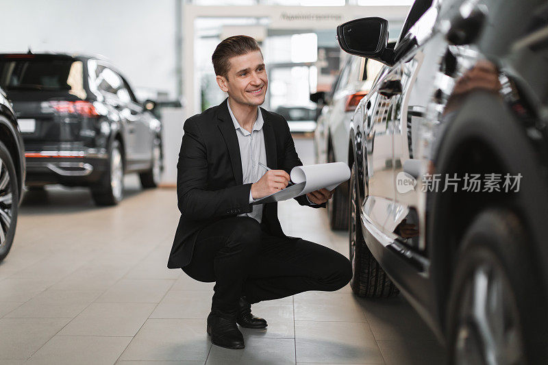 快乐的年轻汽车销售员坐在新车旁边，检查它，在汽车经销商做笔记