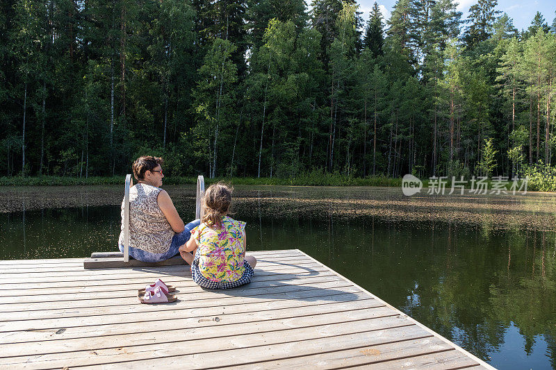 一位77岁的老妇人和一个8岁的白人女孩坐在森林湖的木码头上聊天。奶奶和孙女在度假。