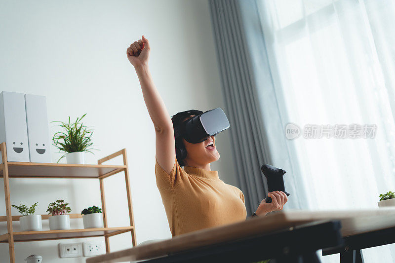 亚洲女性戴着VR头显，拿着操纵杆，在家庭办公室开心地微笑着。