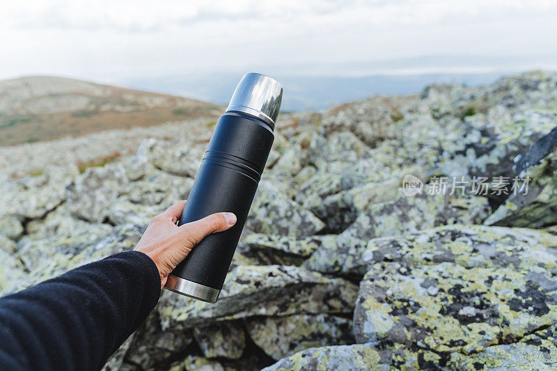 手拿着保温瓶，映衬着群山的背景，黑色的金属保温瓶里装着茶，一位游客在山间用保温瓶喝咖啡，徒步前往难以到达的地方。