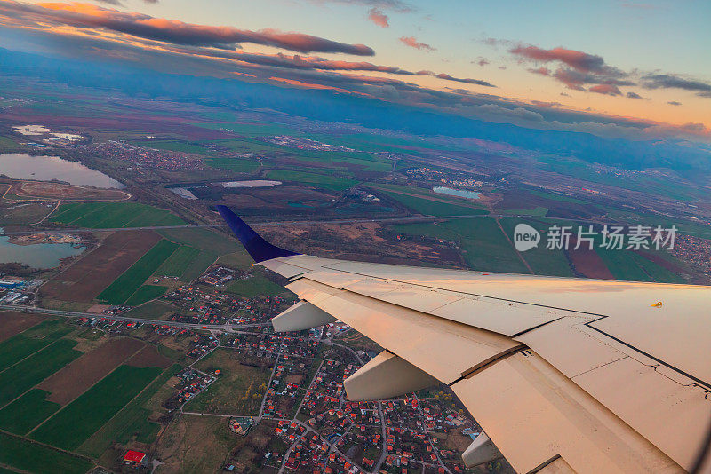 飞机在夕阳的天空中飞过城市和飞机的机翼。从飞机窗口看到的景象。在空中旅行。