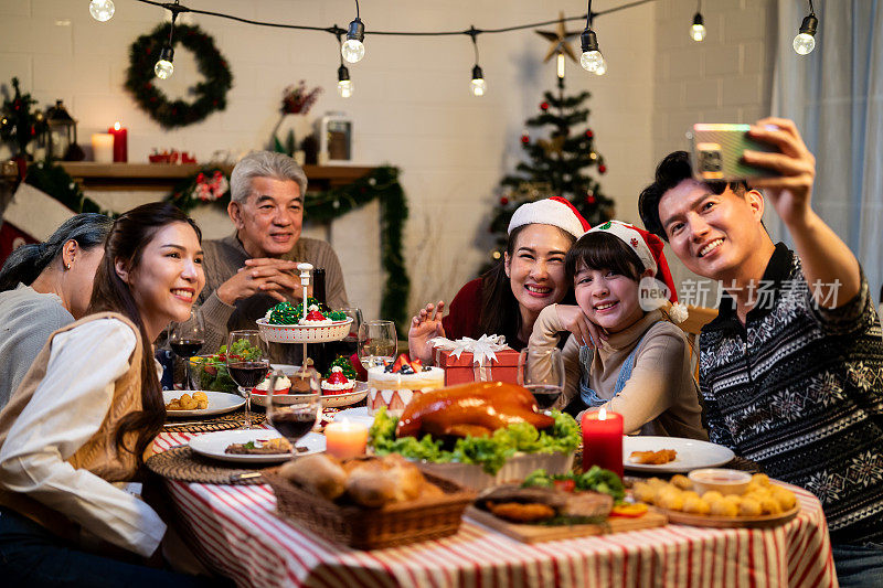 亚洲家庭在一起庆祝圣诞派对时拍照。年轻迷人的孩子女儿和父母在家里的餐桌上吃晚餐，享受节日感恩节、圣诞前夜的美食。