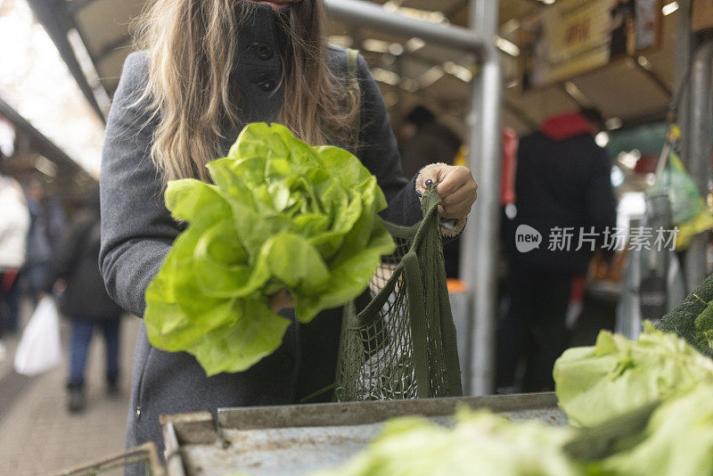 一位妇女在街市上买蔬菜。她把买来的蔬菜放进网袋里