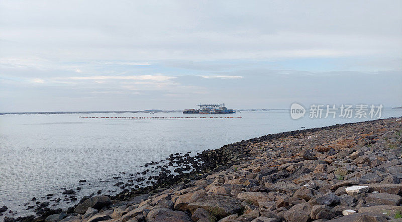 灰色粉砂岩和鹅卵石组成的海岸堤岸，背景是一艘建筑船和一个填埋过程，它制造了几个填海岛屿