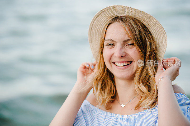 时尚女性特写肖像。快乐的美丽女孩站在沙滩上，享受夏日的假期。戴草帽的金发女郎微笑着走到海边，看着镜头。