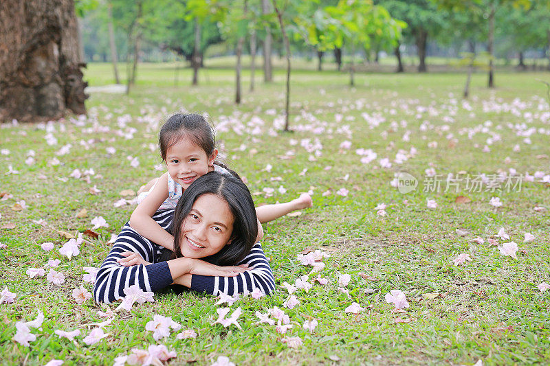 欢快的小女孩依偎着妈妈躺在花园的绿色田野上，粉红色的花已经落满了。幸福美满的家庭。