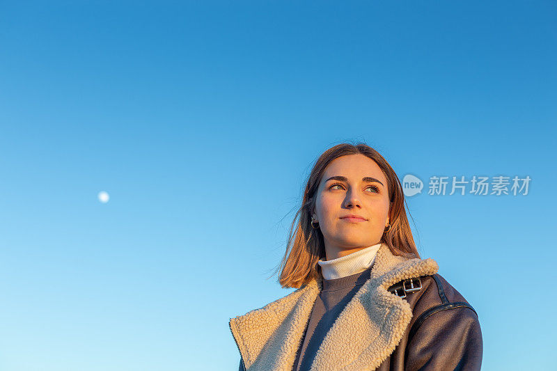 一个美丽的年轻女子望向远方的低角度视图，被落日的光照亮，映衬着蓝天和月亮。