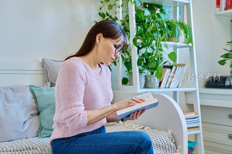 成熟的女人坐在家里的沙发上看纸质书。