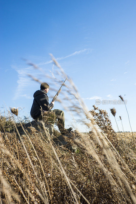 一个猎人拿着猎枪坐在农田边上的岩石堆上。