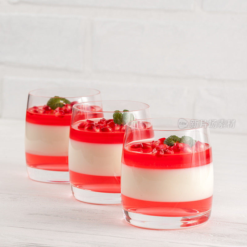 奶油香草意式奶冻配红色果冻在美丽的玻璃杯里，新鲜成熟的石榴在白色的木质背景上。