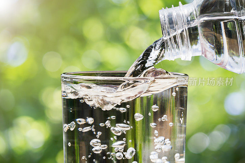 特写水从饮用水瓶流入玻璃上模糊的绿色自然散景背景，健康饮用干净的水的概念