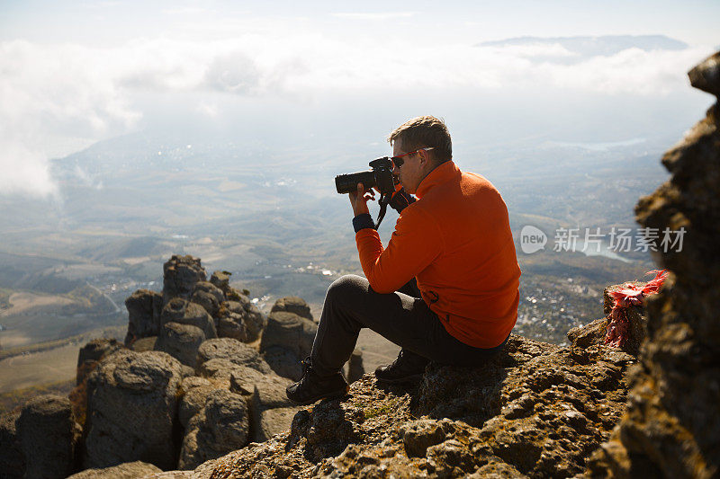年轻的专业旅行家拿着数码单反相机拍摄户外美妙的山景。一个徒步旅行者坐在山顶的一块岩石上，在云层上摄影