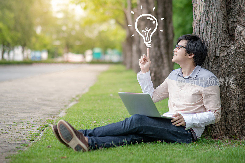 年轻的亚洲人大学学生与眼镜指着灯泡图形插图，而使用笔记本电脑在大学公园学习。创造性思维理念和灵感概念