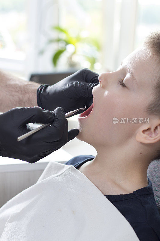 牙医的手上戴着一次性黑色橡胶手套，拿着一把牙科挖土机。这个学龄男孩张着嘴坐在椅子上。医生正在检查男孩的牙齿。