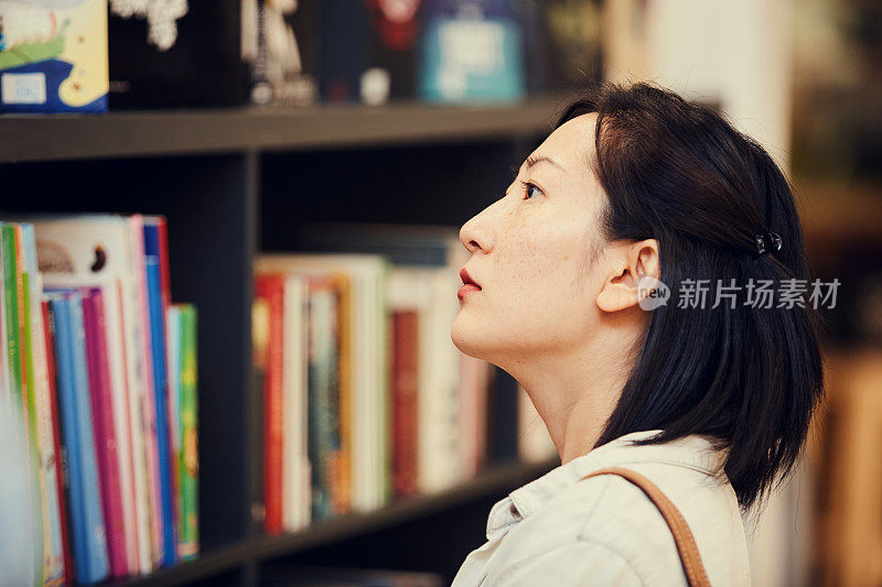 特写严肃沉思的年轻亚洲女人短发站在书店和选择书架上的书