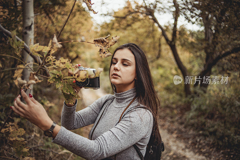 在大自然中摄影——快乐的年轻女子微笑着享受大自然，走过画着秋天颜色的森林，戴着复古的相机拍摄树叶