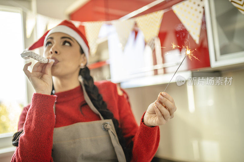 年轻的女士戴着圣诞老人的帽子，吹着喇叭，拿着蜡烛在她的厨房里