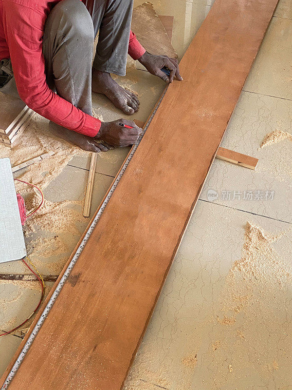 印度勤杂工蹲着，用金属卷尺测量木板，铺着锯末的瓷砖地板