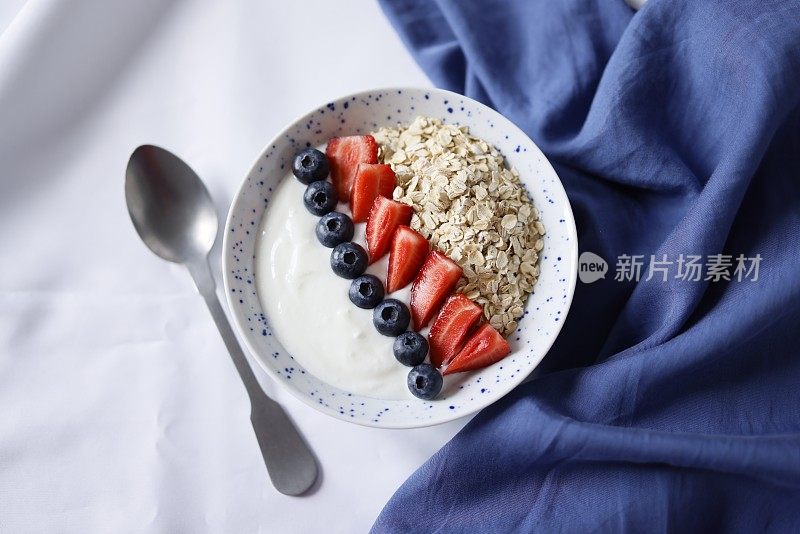 一碗健康早餐，用燕麦酸奶装饰草莓和蓝莓，盘子旁边是一个哑光金属勺和蓝色餐巾，社交媒体趋势早餐，特写