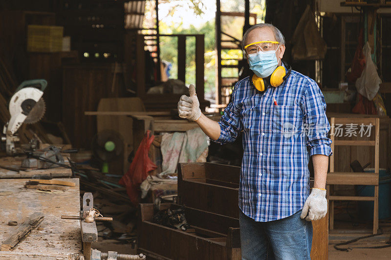老木匠站在木工车间，以家具的工具和产品为背景，竖起大拇指。