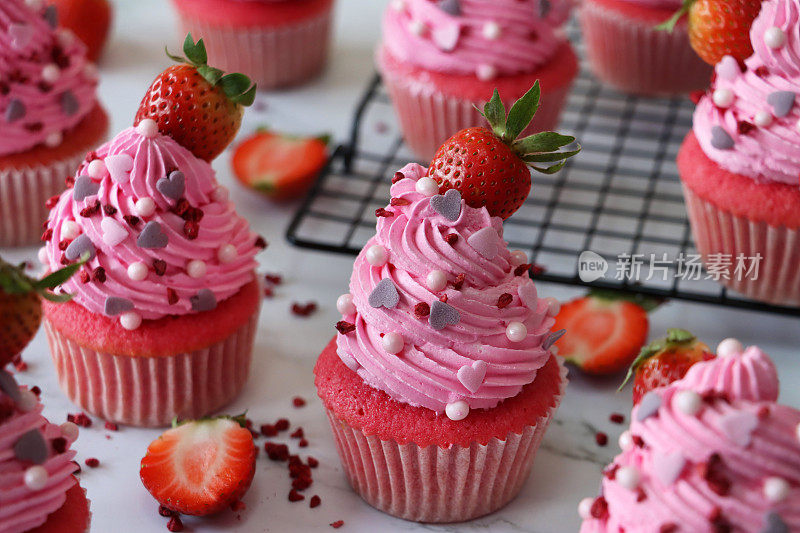 这是一批自制的粉色天鹅绒草莓纸杯蛋糕，放在纸蛋糕盒里，草莓放在粉色奶油糖霜上，糖精做成心形和球形，放在金属冷却架上，关注前景