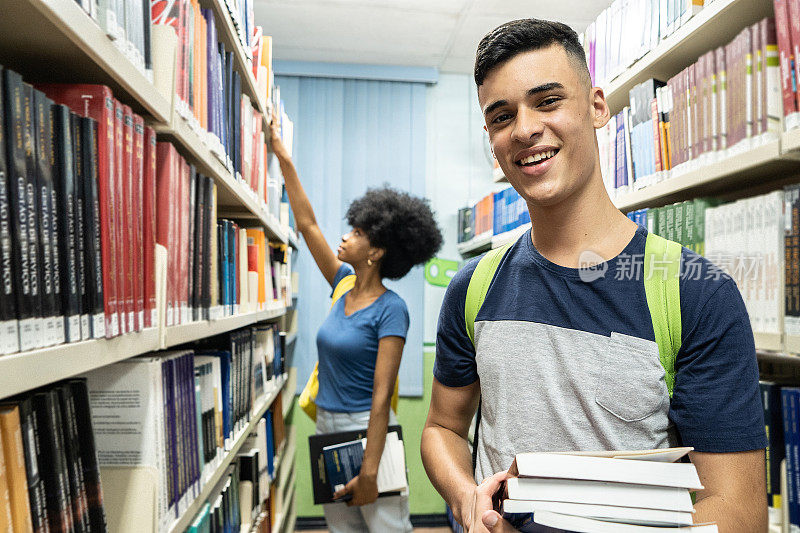 一个十几岁的男孩在图书馆里拿着一些书的肖像，背景是大学