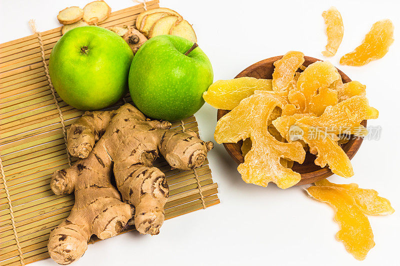 干姜片和糖放在木碗里，新鲜的姜根和绿苹果在白色的背景上。生姜产品具有复制空间。