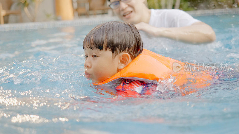 亚裔家庭，父亲在教游泳，和儿子在游泳池里玩水。教练教学生游泳，在假期里开心快乐。孩子们喜欢暑假的各种活动。