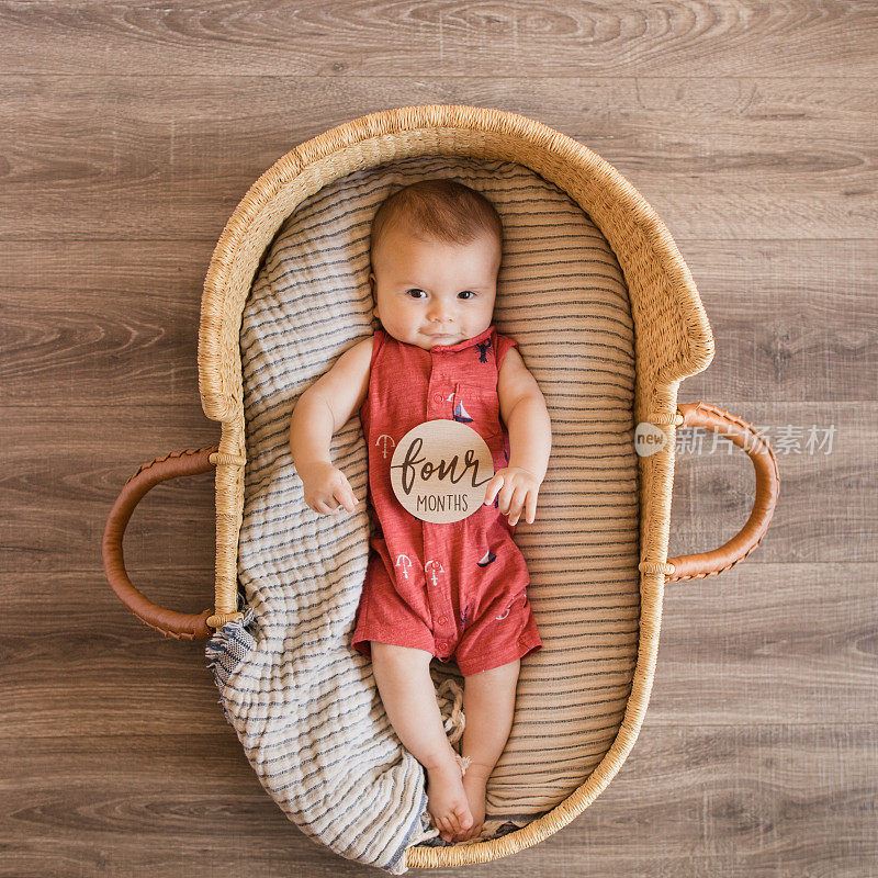 一个4个月大的小男孩穿着一件红色的航海服，躺在一个舒适的奶油色条纹棉毯上，在海草摩西篮子