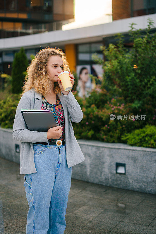 一头卷发的白人女生在去大学的路上喝着早上的咖啡，手里拿着笔记本电脑。生活方式，大学和人的概念。好看。