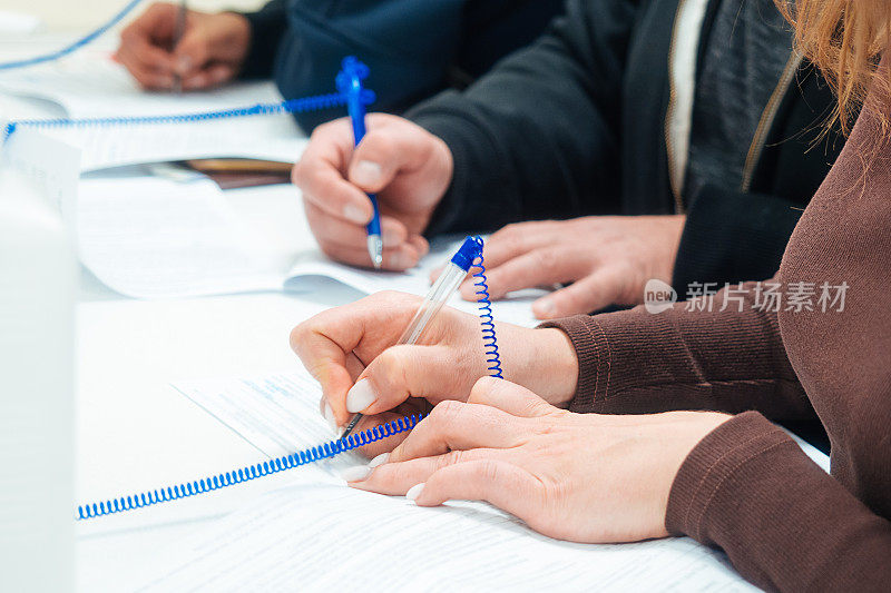 妇女在桌子上填写空白申请表或签署合同。手里拿着近距离的自来水笔。几个人同时用笔在纸上填写数据。