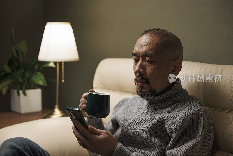 亚洲中年男子坐在沙发上，一边喝咖啡一边看手机