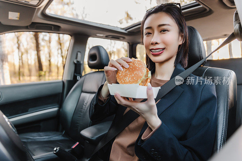 亚洲女人在车里休息，吃汉堡。一个年轻成功快乐的女人正在开车。